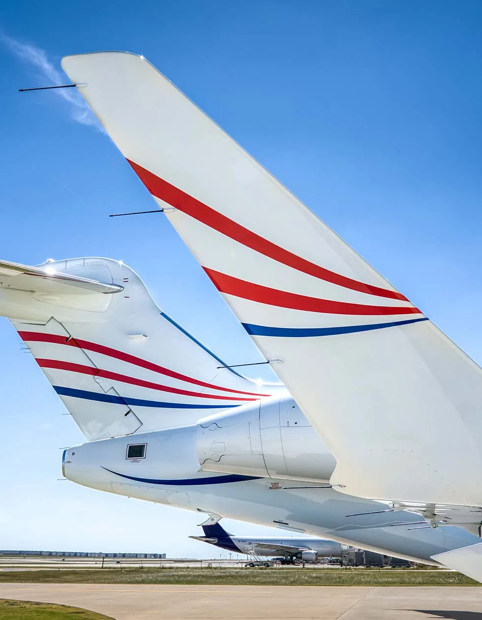 The tails of several white business jets with red and blue stripe paint schemes are parked on an airfield under a clear blue sky.