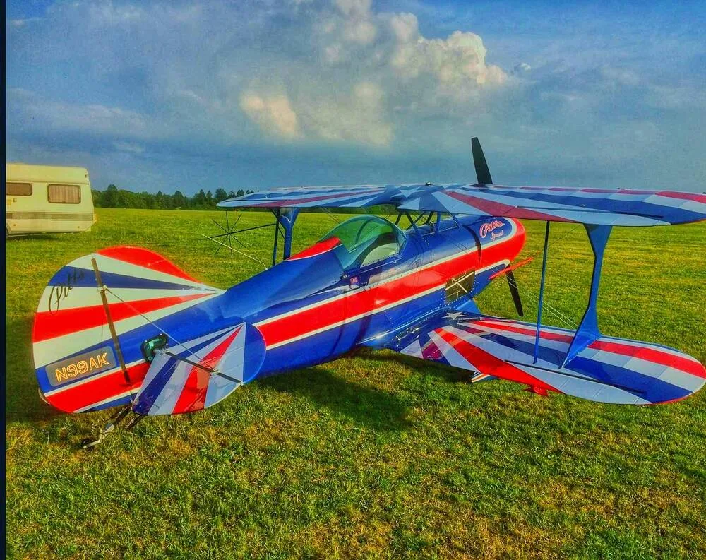 A red, white, and blue Pitts Special biplane, N99AK, rests on a grassy field with a camper in the background.