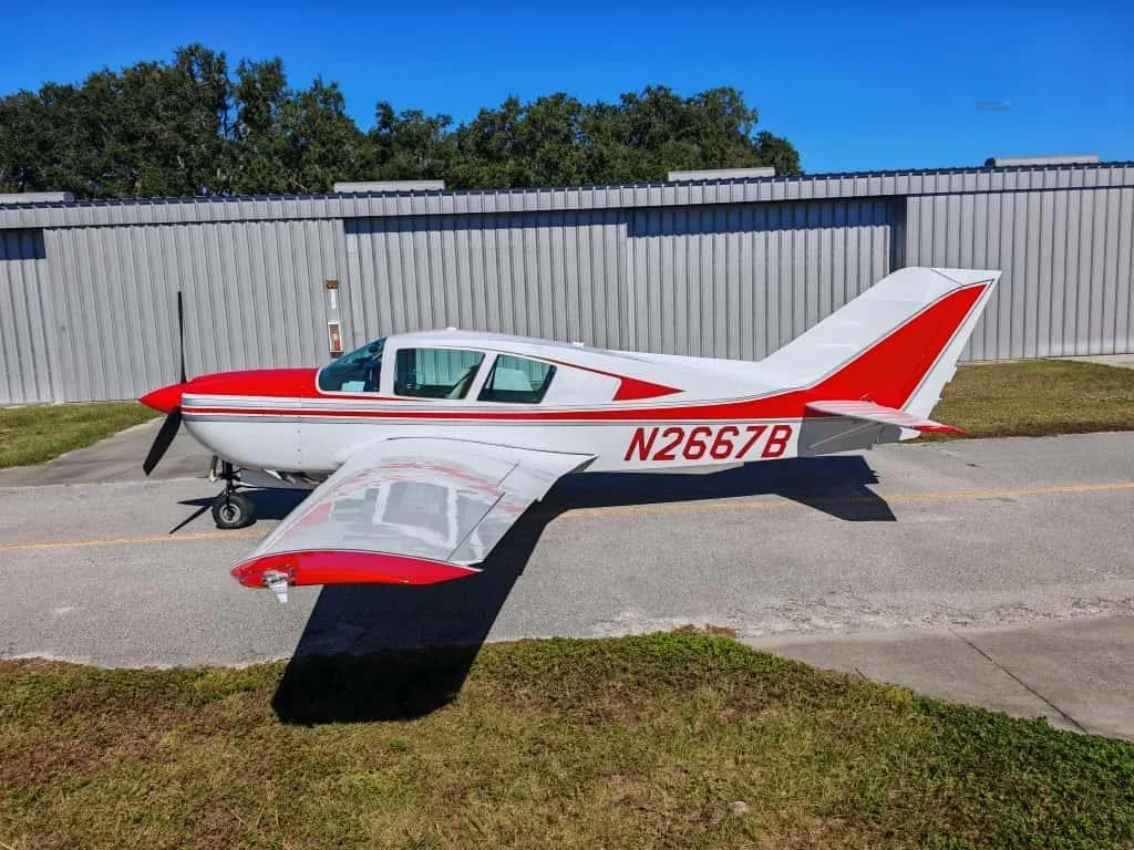 A white and red single-engine propeller aircraft (N2667B) is parked on an asphalt ramp in front of a metal hangar under a clear blue sky.