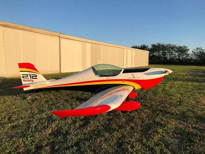 A white, red, and yellow single-engine aircraft with a bubble canopy sits on a grassy field at sunset.