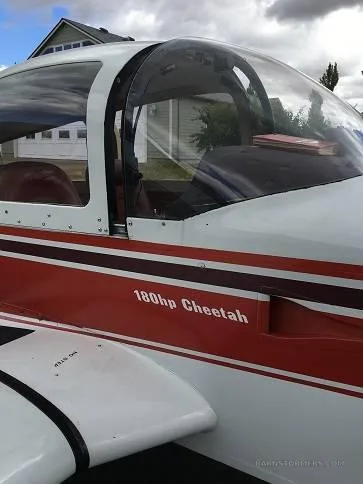 A close-up of the cockpit canopy and fuselage of the white and red Grumman American AA-5, showing '180hp Cheetah' text.