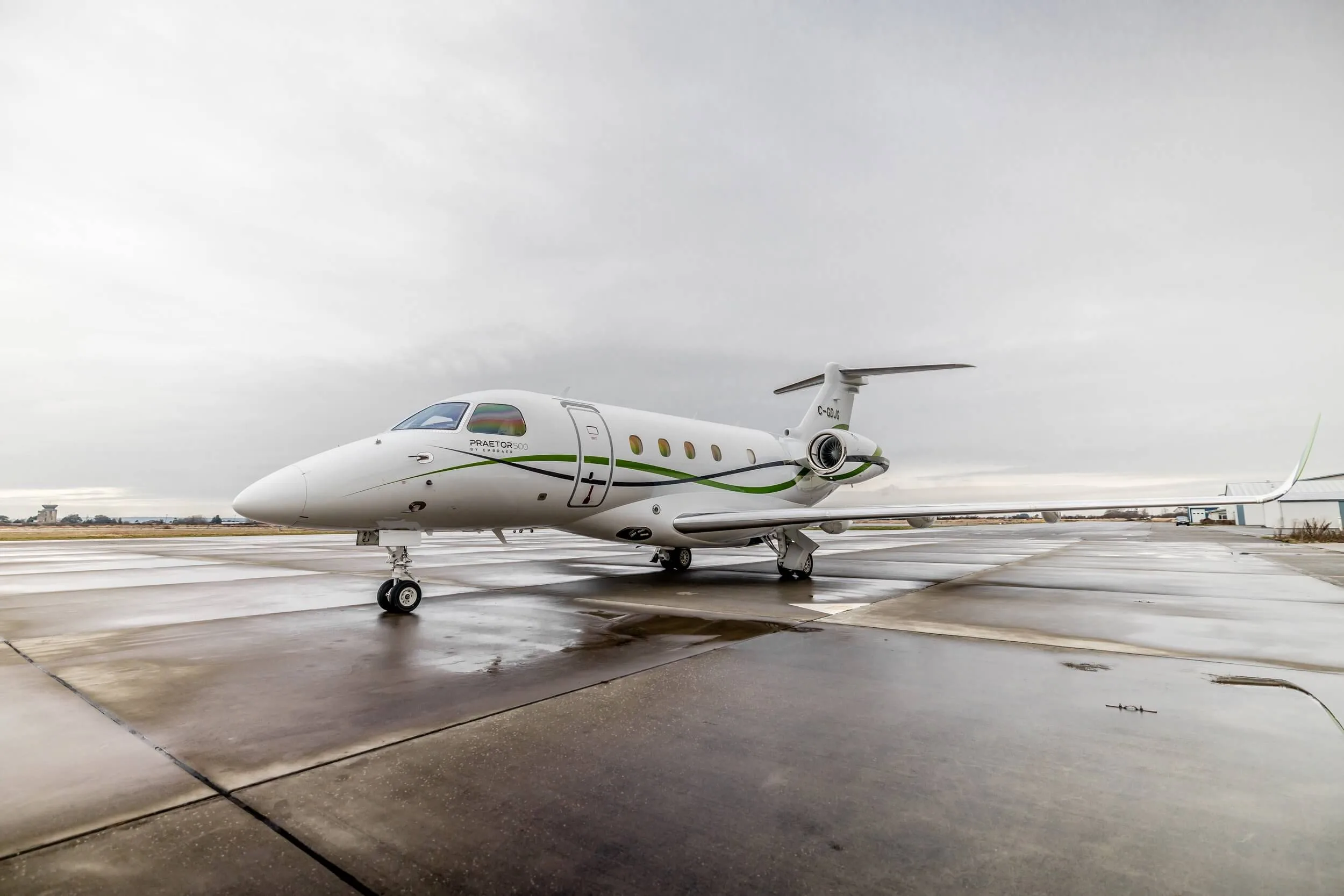 A white Embraer Praetor business jet with green and black stripes is parked on a wet tarmac under an overcast sky.