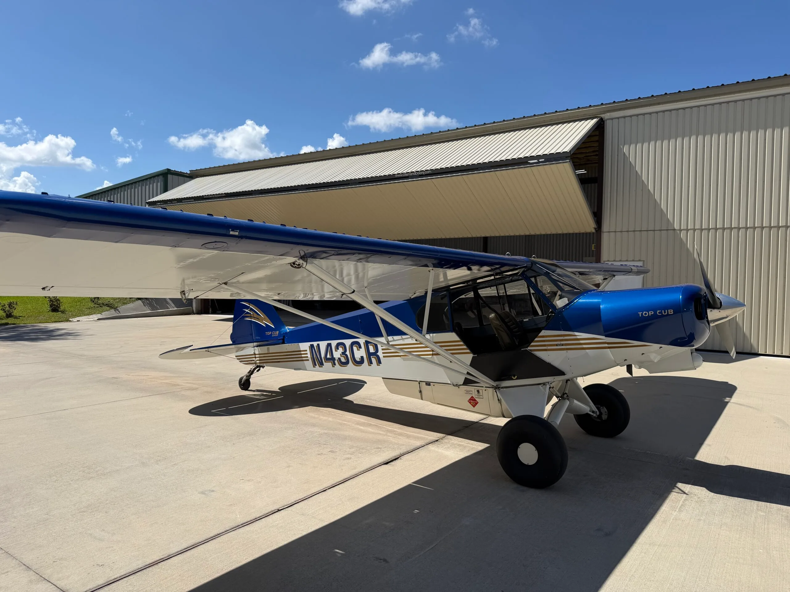 A blue and white Top Cub aircraft with gold stripes, registration N43CR, parked on a concrete tarmac next to a hangar under a blue sky.