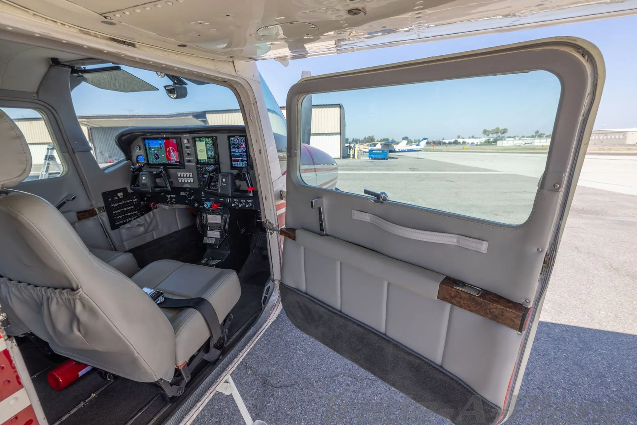 The cockpit and front cabin of a Cessna 210 Centurion aircraft, showing the instrument panel and gray seats, with the door open to the tarmac.