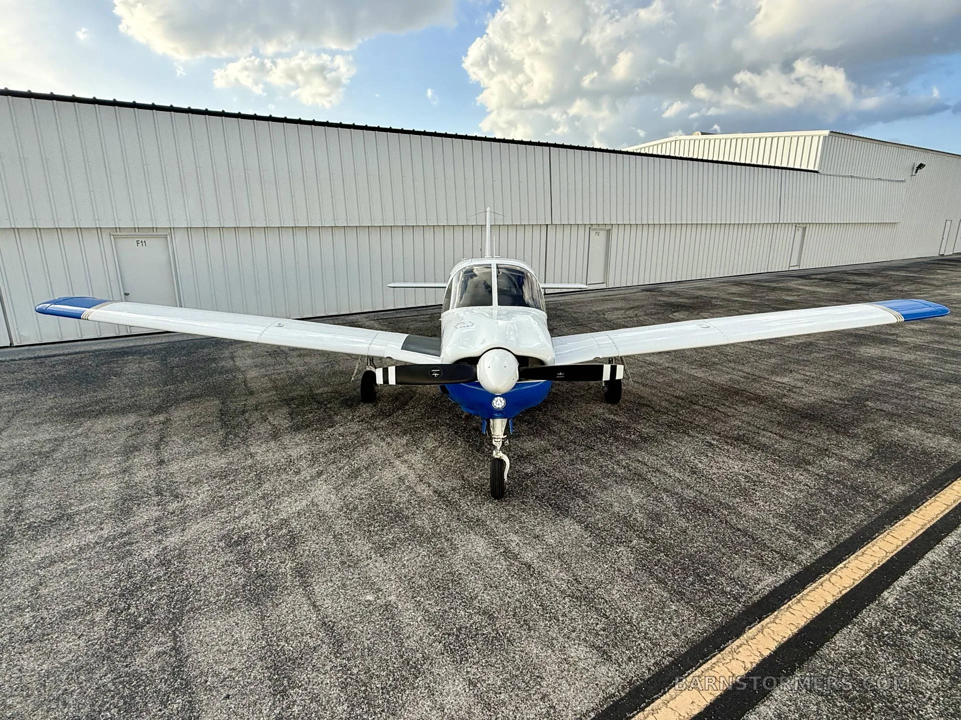 A front view of a white and blue Piper Cherokee aircraft (N338PA) parked on an asphalt ramp in front of a hangar.