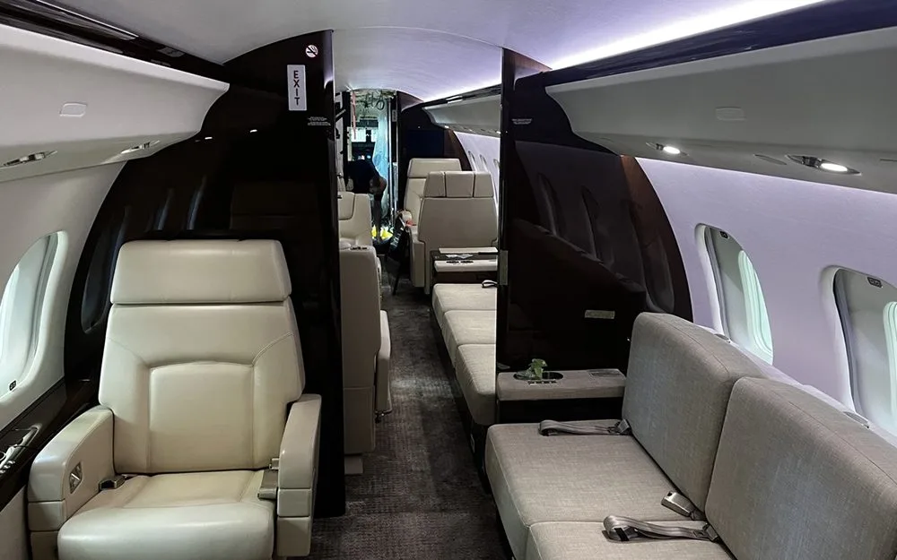 The interior of a private jet cabin showing cream-colored seats and a sofa, with dark wood paneling.