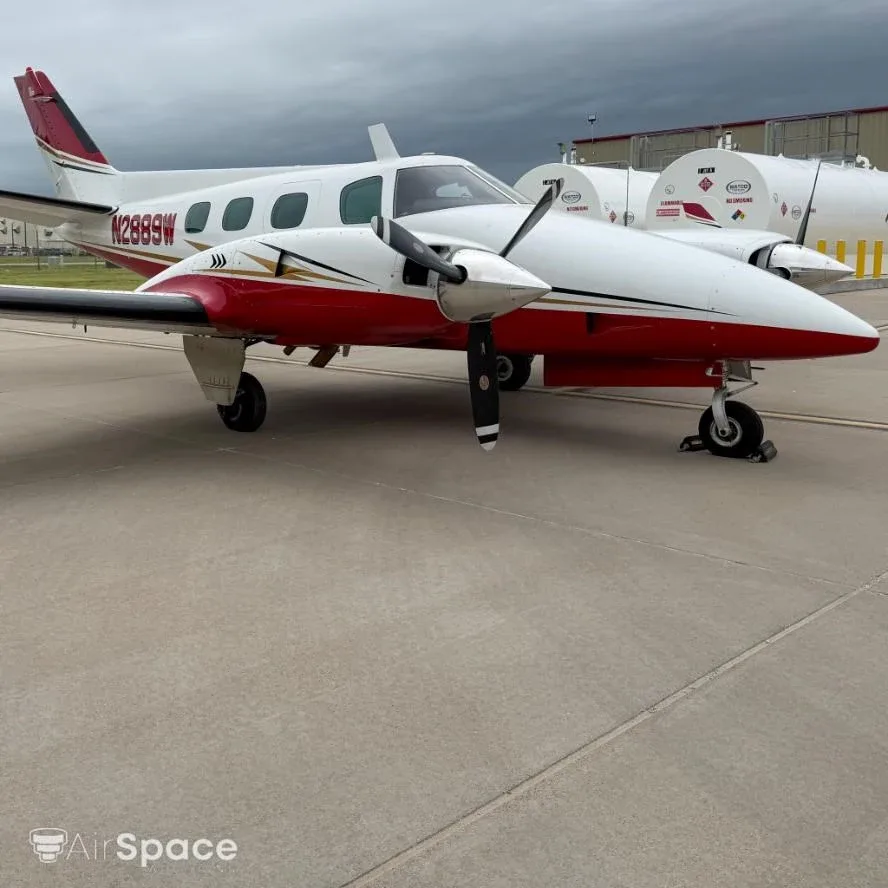 A white and red Beechcraft Duke N2889W parked on a tarmac with fuel tanks in the background under a cloudy sky.