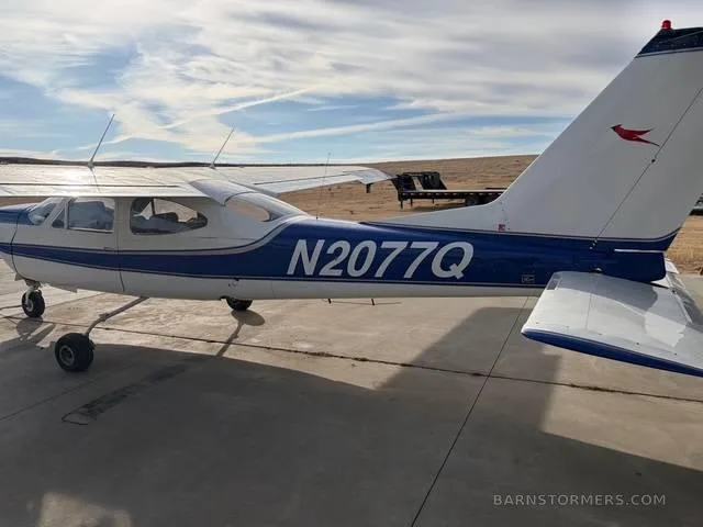 A white and blue Cessna aircraft with tail number N2077Q parked on a tarmac, viewed from the rear right.