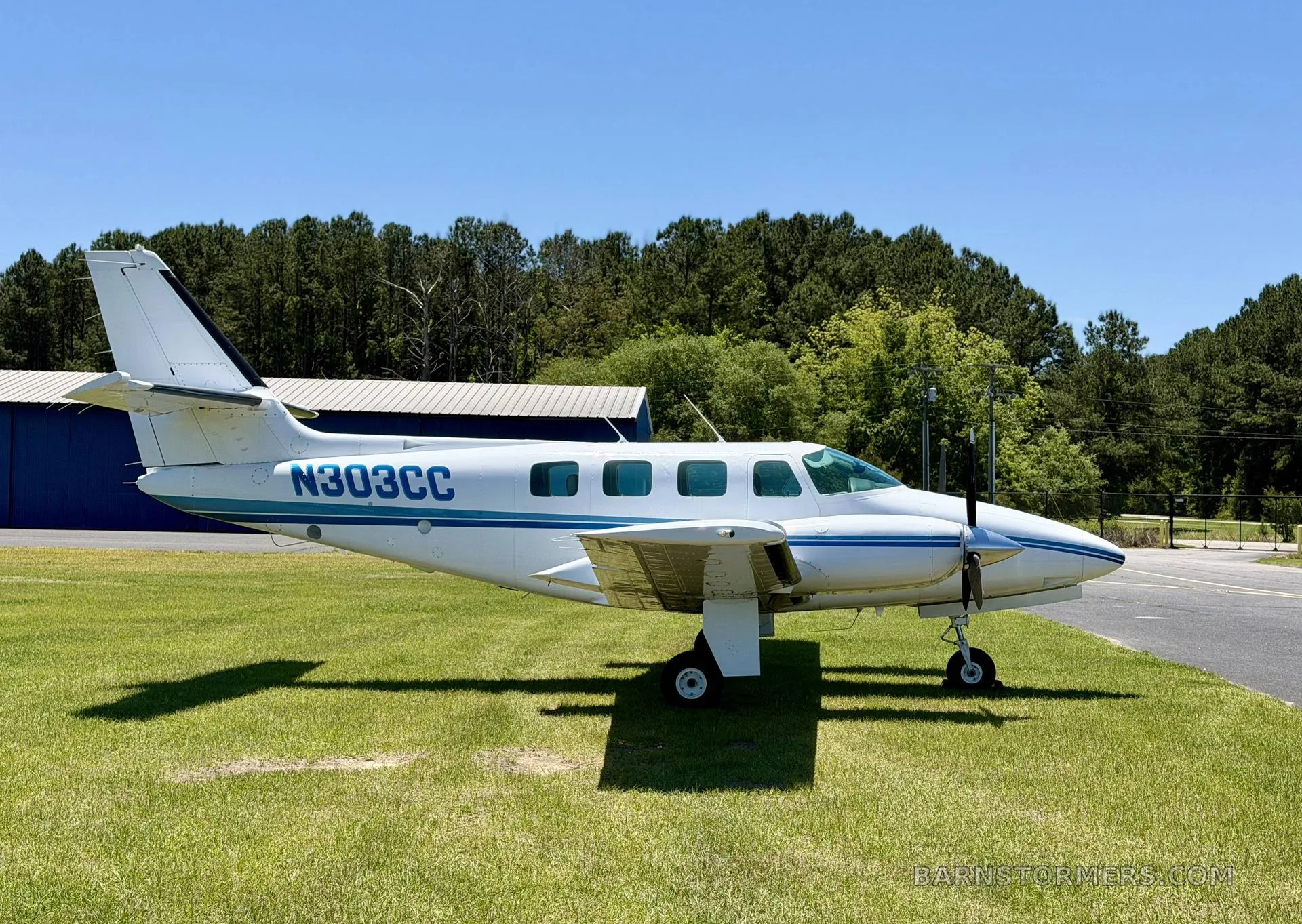 A white twin-engine propeller aircraft with blue stripes (N303CC) is parked on a grassy field with a blue hangar in the background.