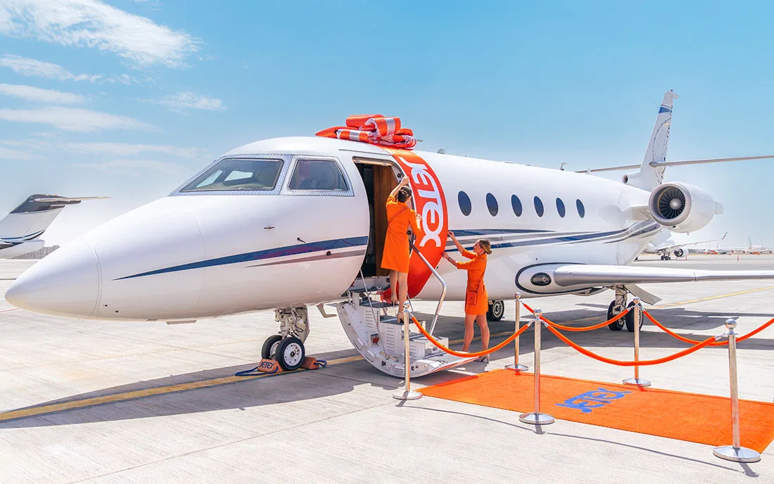 A white business jet with an orange 'Jetex' ribbon and steps down, with two people in orange uniforms near the entrance on a sunny tarmac.