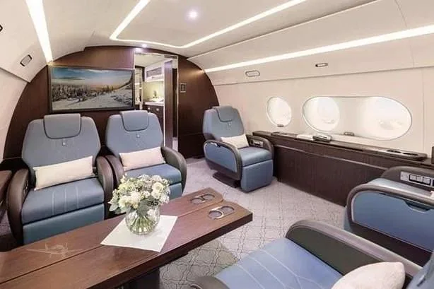 A spacious aircraft cabin featuring blue leather seats, a dark wood table with flowers, and a large screen displaying a snowy landscape, viewed from a rear perspective.