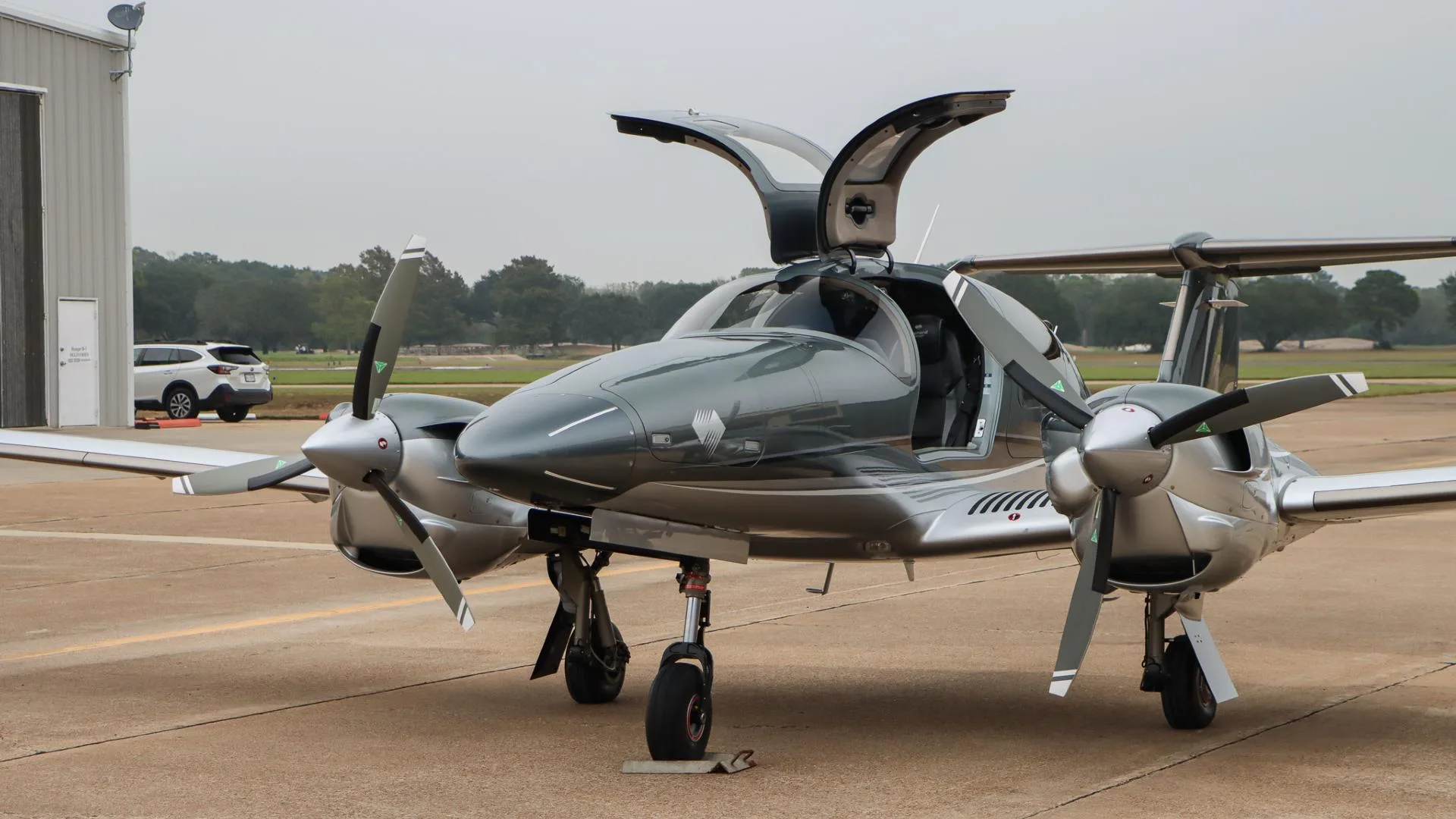 A front-side view of the gray and silver Diamond DA62 aircraft with its gull-wing doors open, parked on an airport tarmac.