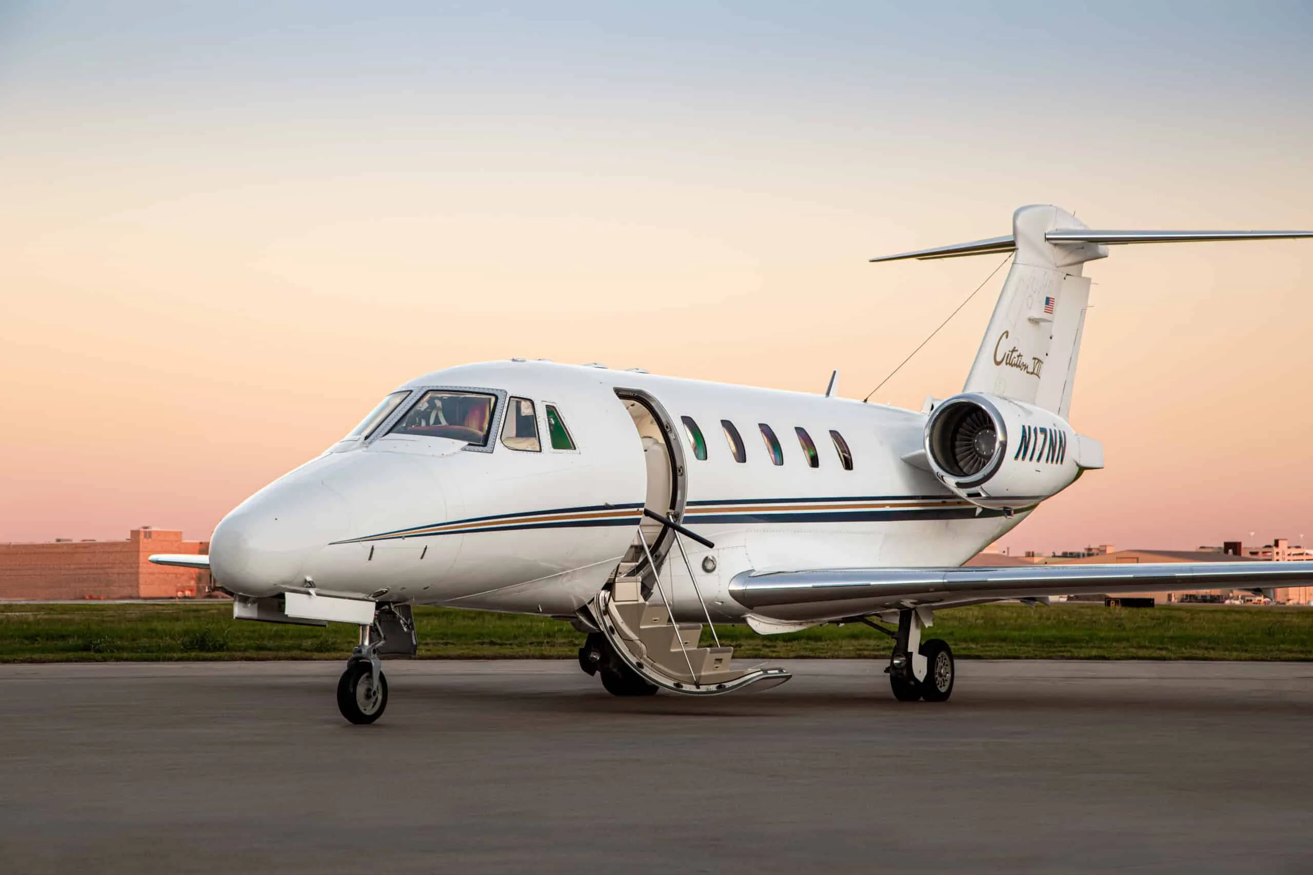 A white Cessna Citation III business jet with blue and gold stripes, registration N77NN, parked on a tarmac with its door open at sunset.