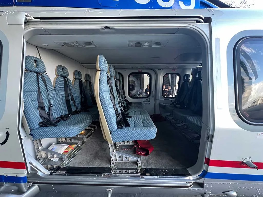The open side door of a blue and white helicopter revealing rows of blue passenger seats inside a hangar.
