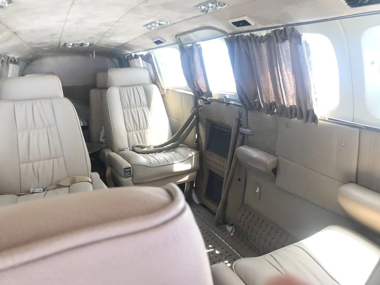 The tan leather interior cabin of a small aircraft with multiple seats and windows with brown curtains.