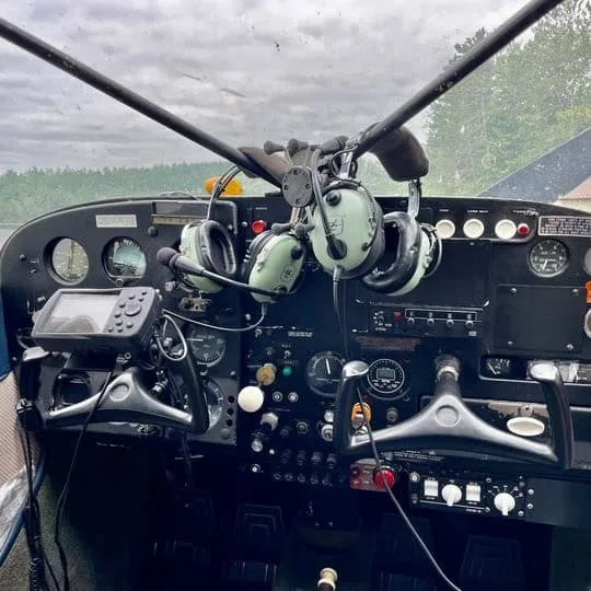 The cockpit of a floatplane shows the instrument panel, controls, and two headsets hanging.