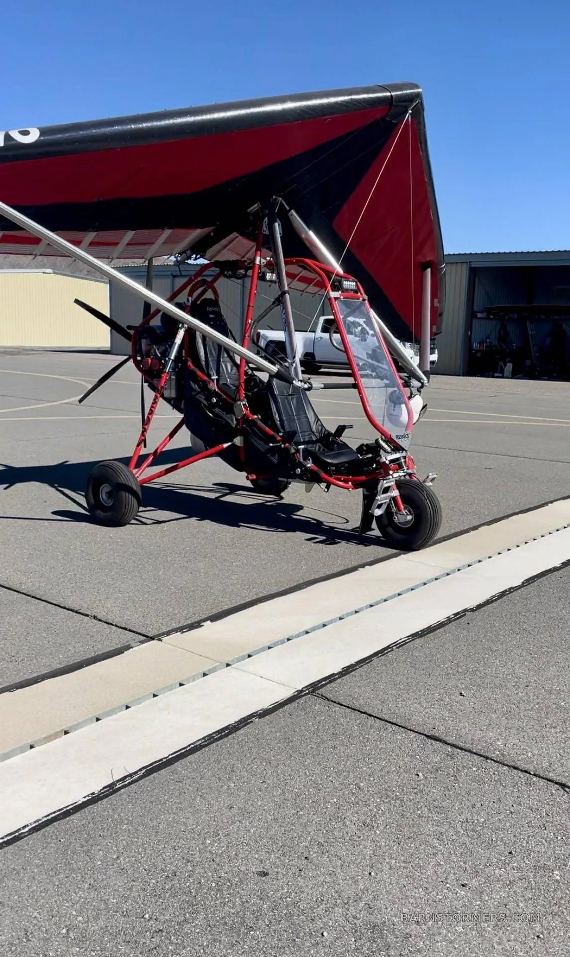 A red and black trike ultralight aircraft with a 'REVOLT' logo, parked on an airport tarmac next to a hangar.