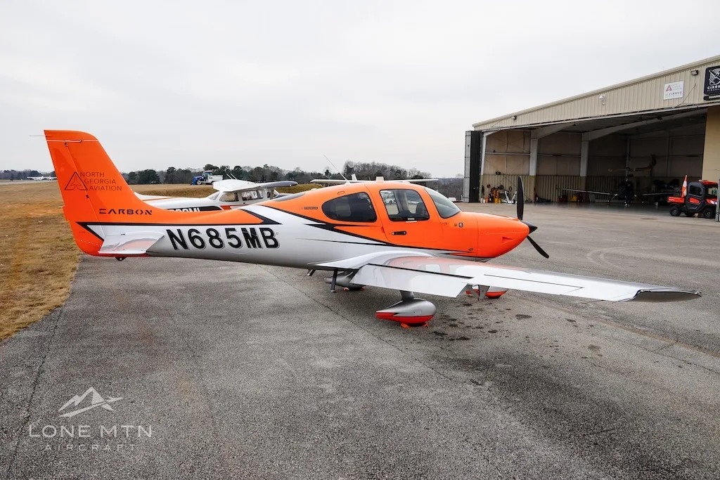 The orange and silver Cirrus SR20 aircraft N685MB parked on an airfield with hangars in the background.