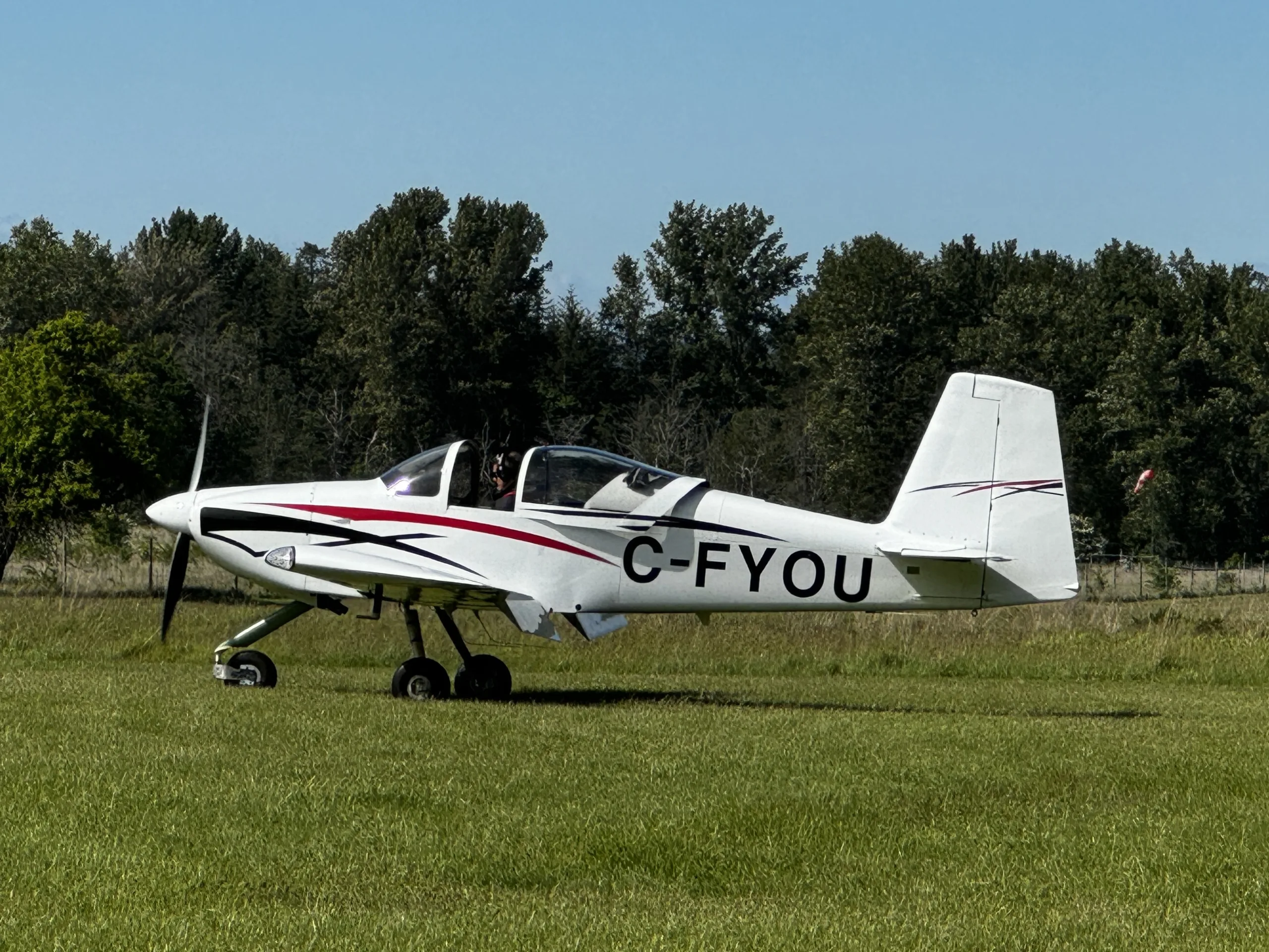 A white single-engine aircraft with red and black stripes, registration C-FYOU, on a grassy field with trees in the background under a clear sky.