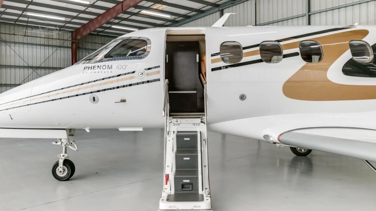 The front section of a white Embraer Phenom 100 jet with gold and black stripes, registration N100NV, showing the open entry door with stairs in a hangar.