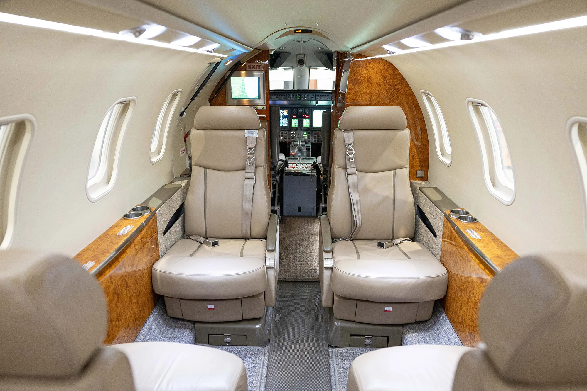 The luxurious interior cabin of a private jet with two tan leather seats in the foreground and the cockpit visible beyond.