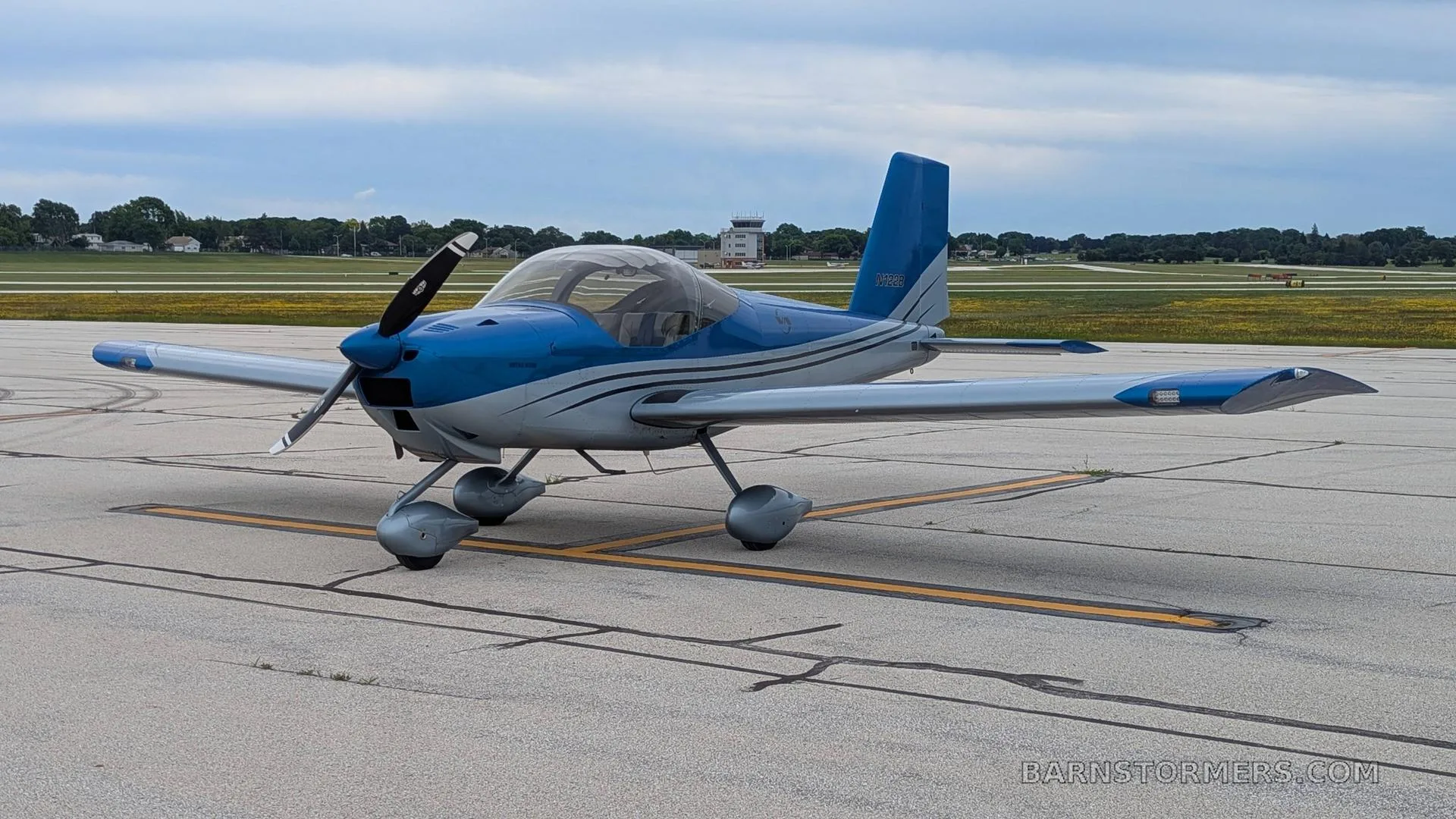 A blue and silver single-engine propeller aircraft, registration N122B, parked on an airfield with a control tower in the distance.