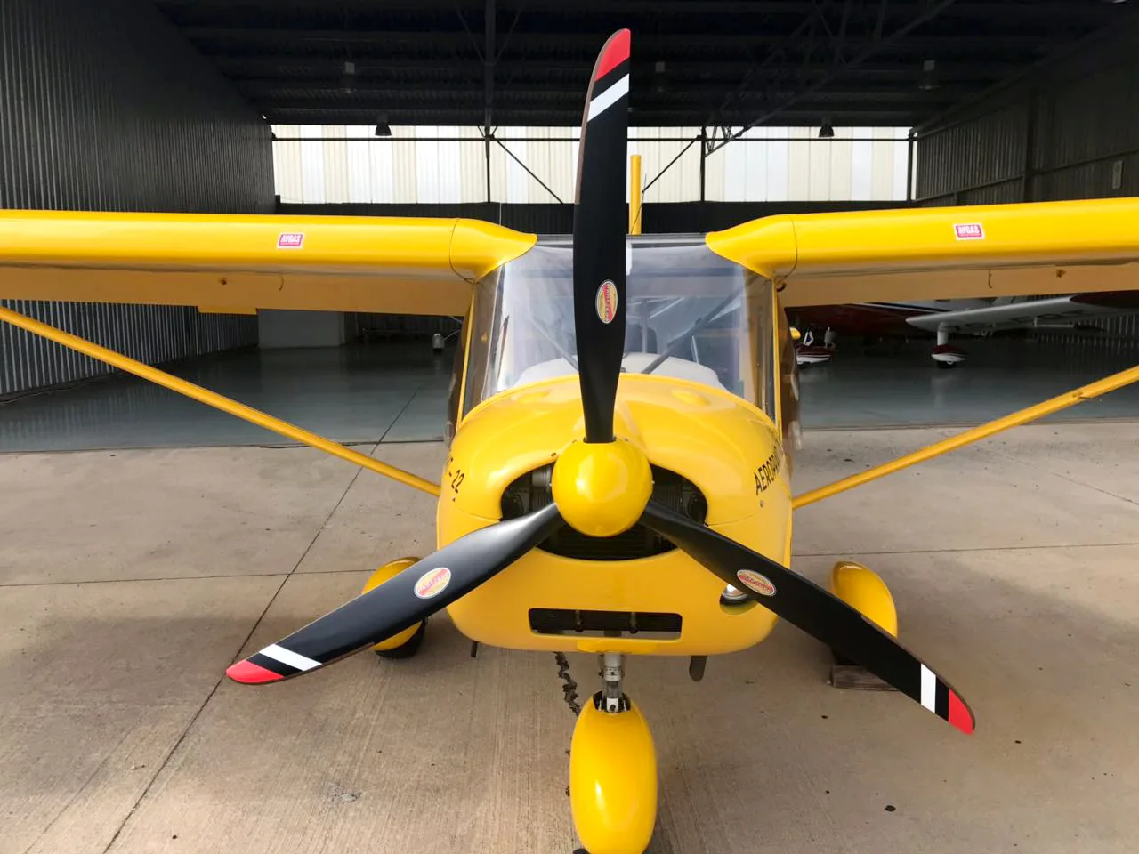 A head-on view of a yellow Aeroprakt-22 aircraft with a black propeller, parked inside a hangar.