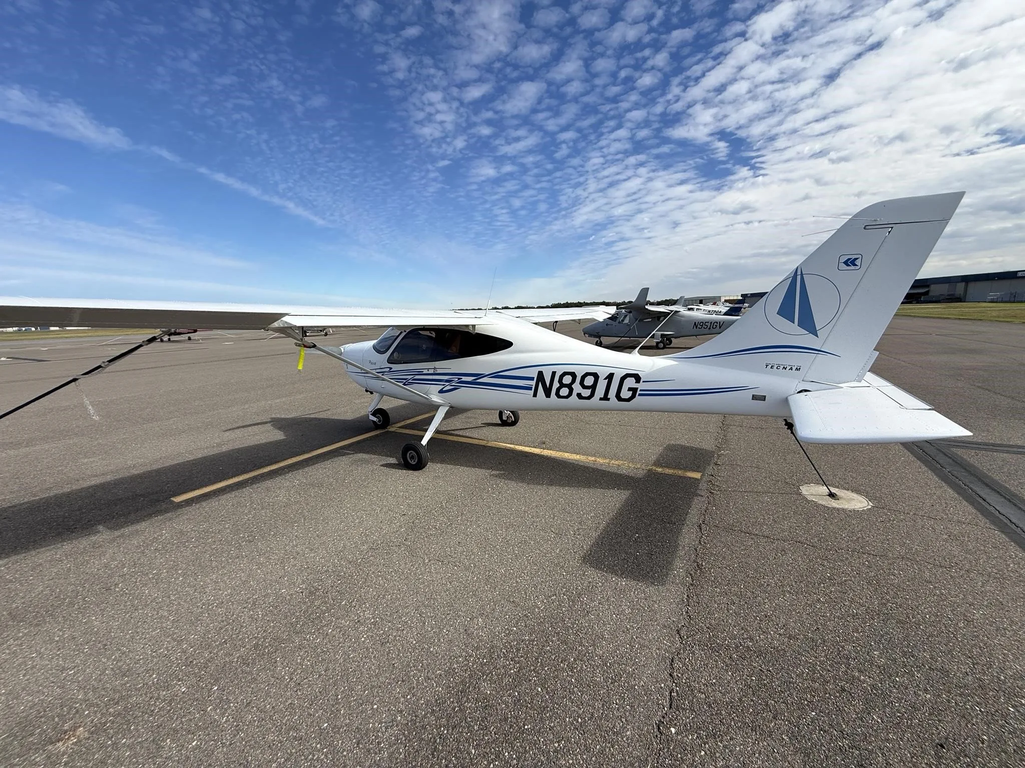 A white Tecnam P2008 aircraft with blue stripes and N891G registration, viewed from the rear-left on an airport tarmac.