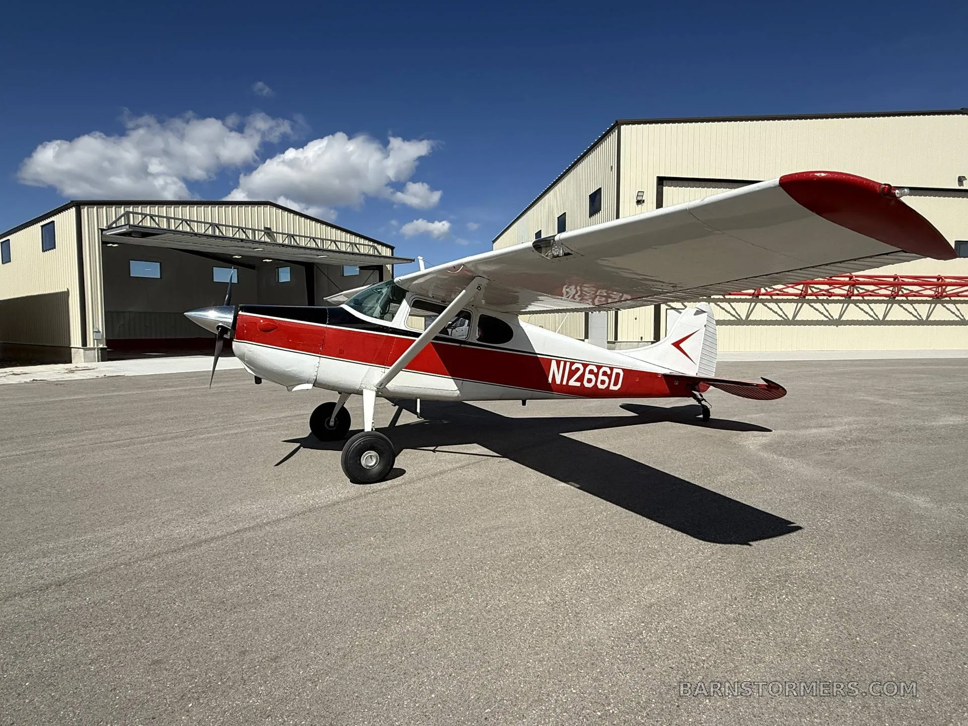 A red and white Cessna 170B (N1266D) is parked on an airfield, viewed from the side-right, with hangars in the background.