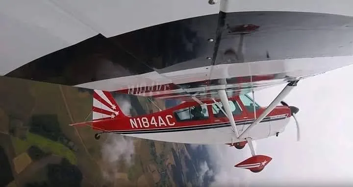 A red and white Super Decathlon aircraft (N184AC) is seen in flight from below, with land visible in the background.