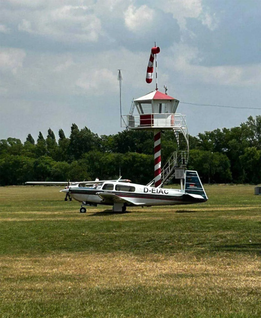 A Mooney M20J aircraft with a silver, blue, and red paint scheme parked on a grassy field with a small red and white striped control tower in the background.