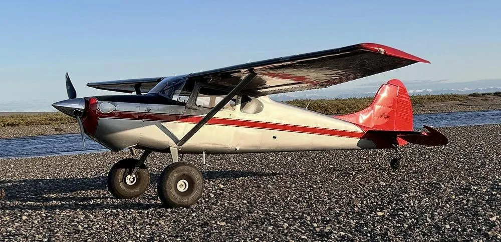 A silver and red Cessna 170B aircraft with large tires parked on a gravel bar next to a river.