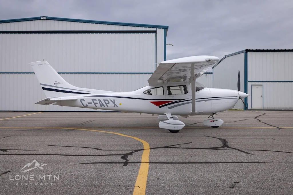 A white Cessna 182 Skylane with grey and blue stripes, registration C-FAPX, parked on an asphalt ramp in front of hangars.
