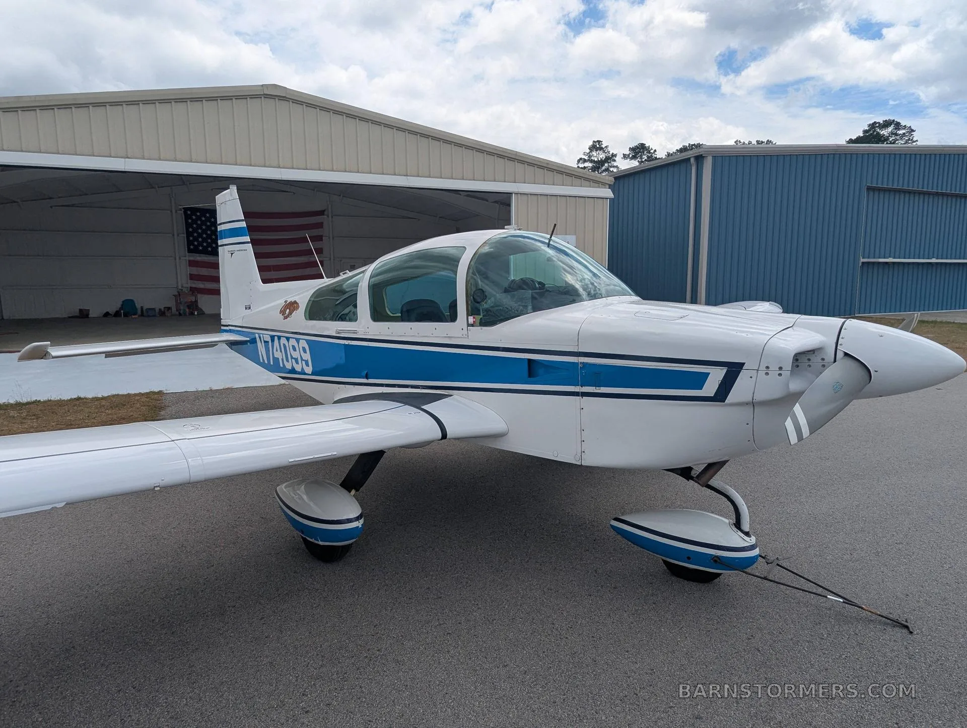 A white and blue Grumman AA-5 aircraft with registration N74099 parked in front of an open hangar with an American flag inside.