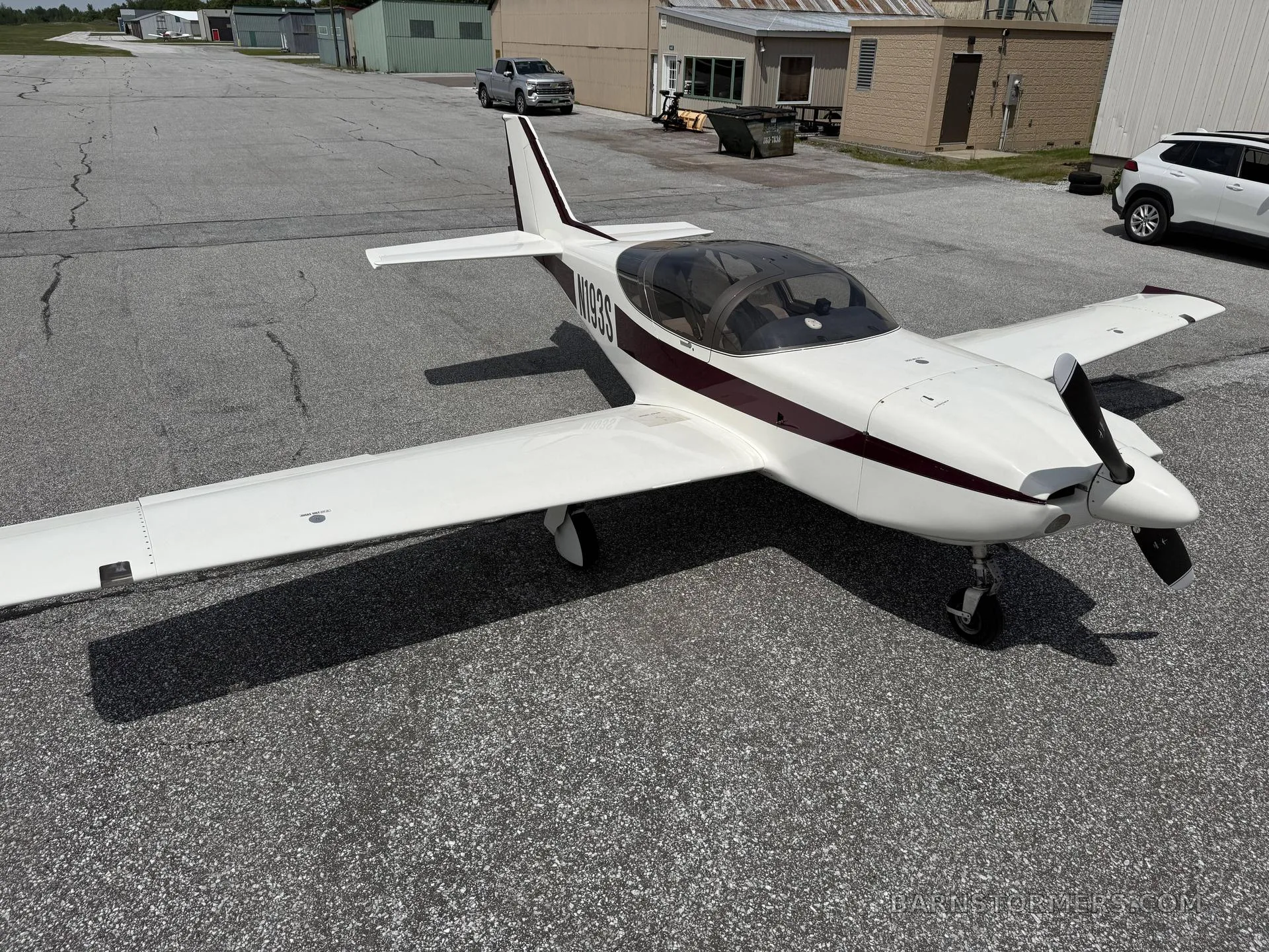 A white and maroon single-engine aircraft (N193S) with a bubble canopy is parked on an asphalt surface with hangars in the background.