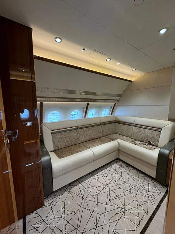 An L-shaped sofa with light upholstery and dark trim is set against the windows in a private jet cabin.