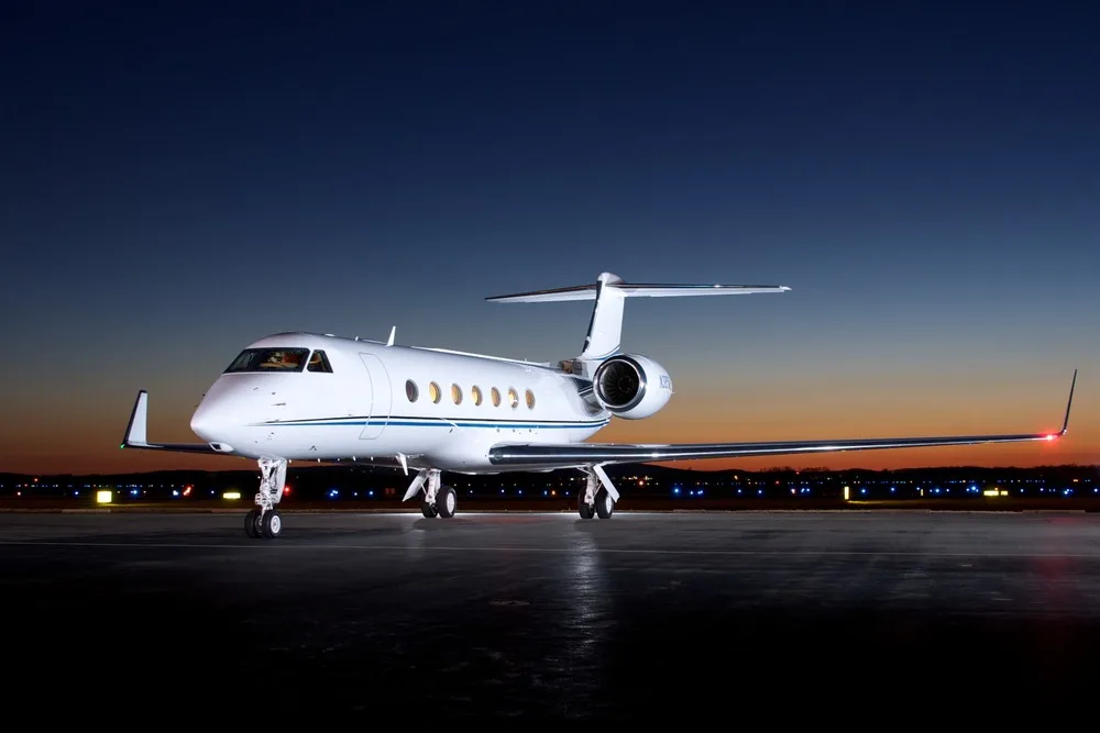 A white Gulfstream G450 business jet with a blue stripe on the tarmac at dusk, viewed from a front-side angle.