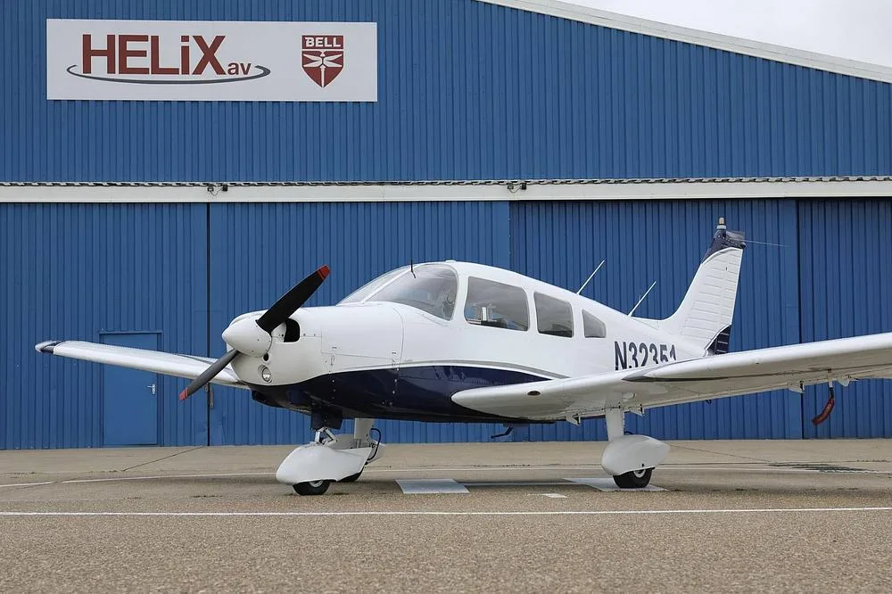 A white and blue Piper PA-28 aircraft with registration N32351 parked on an airfield next to a blue hangar.