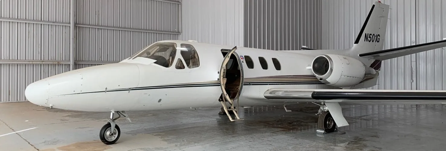 A white business jet with a black and gold stripe, registration N501G, is parked in a hangar with its main door open.