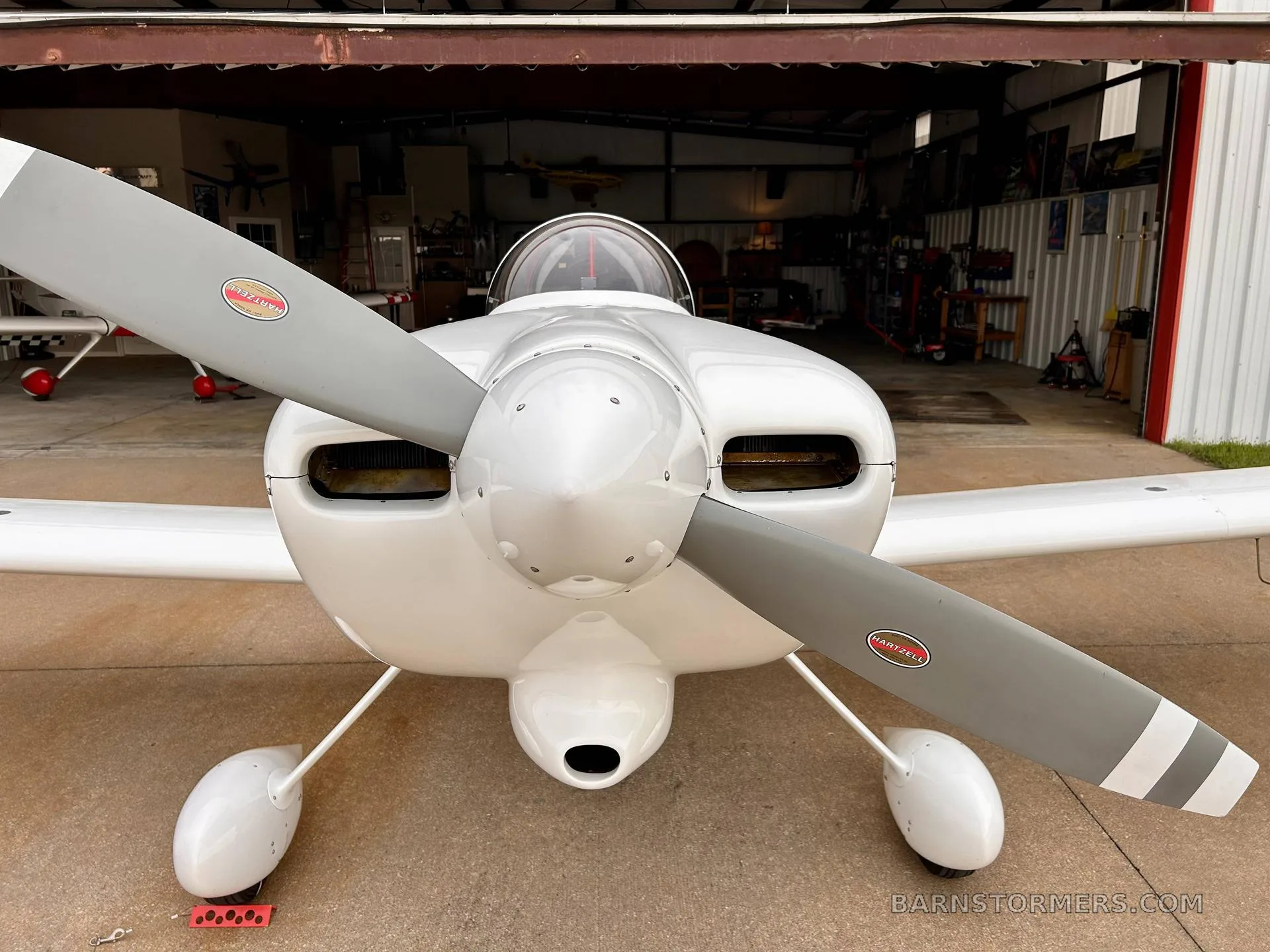 A white single-engine aircraft with a gray propeller, viewed head-on, parked in front of an open hangar.