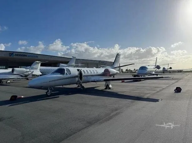 A white business jet is parked on an airport tarmac with other aircraft and a hangar in the background under a blue sky.