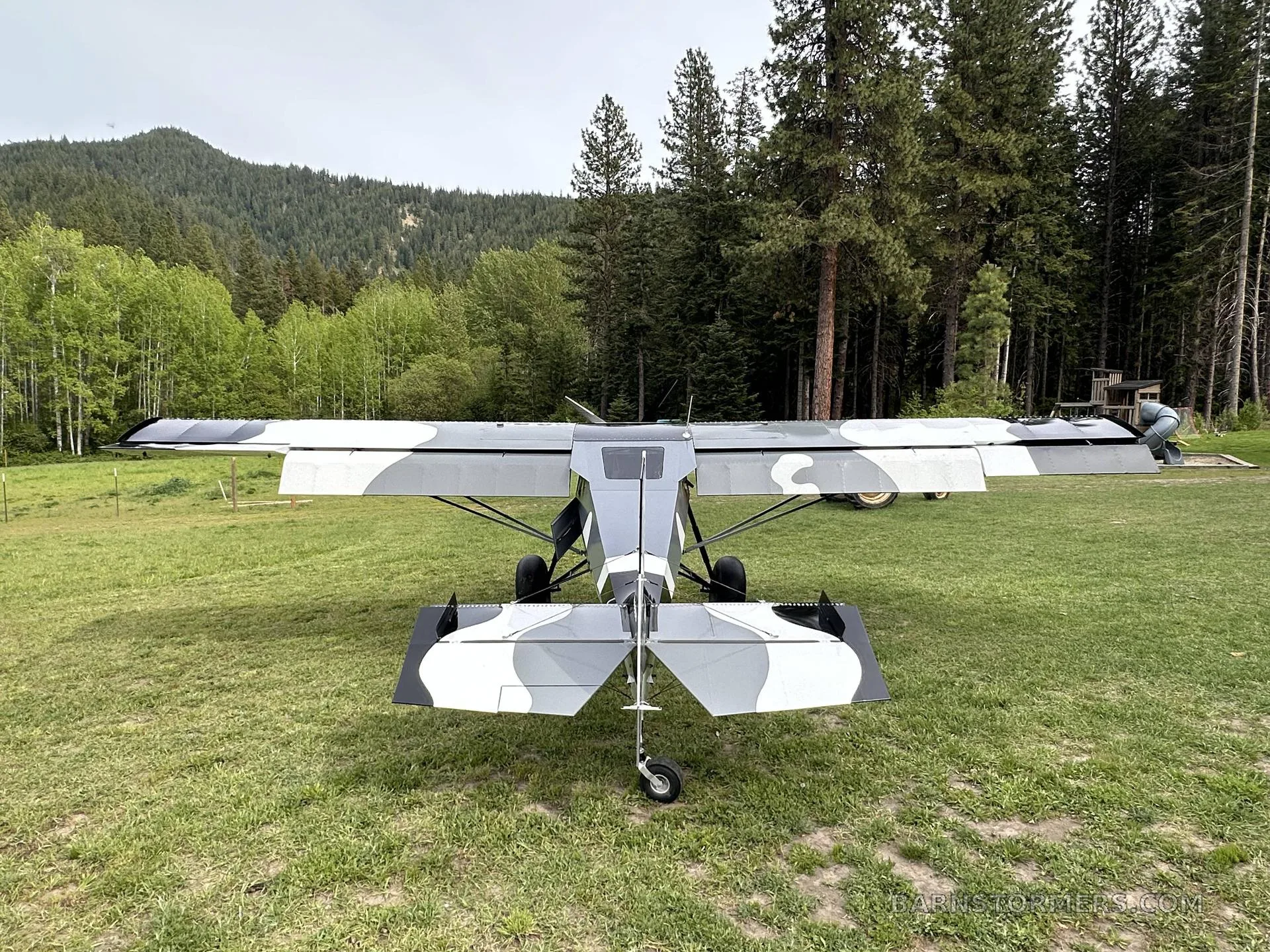 A black, white, and gray camouflage STOL aircraft with large tundra tires is viewed from directly behind, parked on a grassy field.