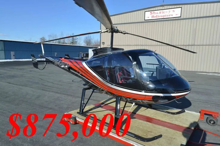 A black and orange Bell 505 Jet Ranger X helicopter with registration N1480L is parked on a tarmac in front of a hangar.