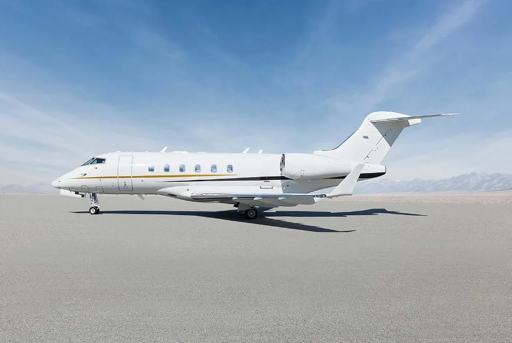 A white Bombardier Challenger 350 business jet with gold and black accent stripes parked on a tarmac, viewed from the side.