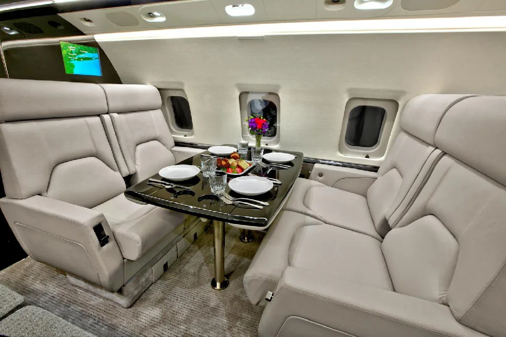 A section of the private jet cabin with two sets of facing light gray seats and a dark table set with plates and fruit.