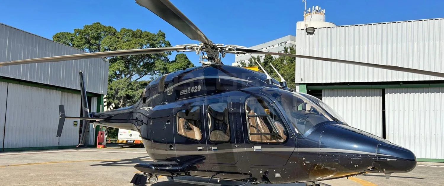 A dark blue Bell 429 helicopter with tan interior visible, parked on an airfield next to a hangar.