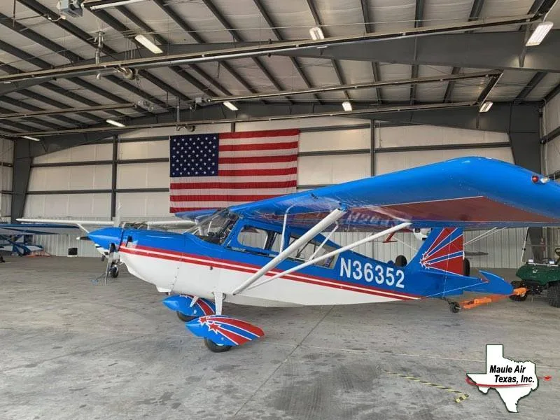 A blue and white Maule M-7-235B aircraft with registration N36352 is parked in a hangar with an American flag in the background.