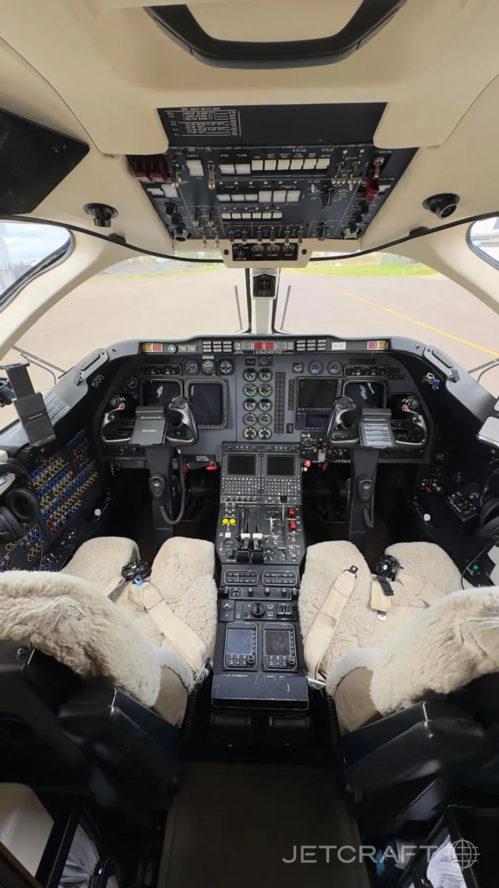 The cockpit of a private jet shows a detailed instrument panel, multiple screens, and two pilot seats with sheepskin covers.