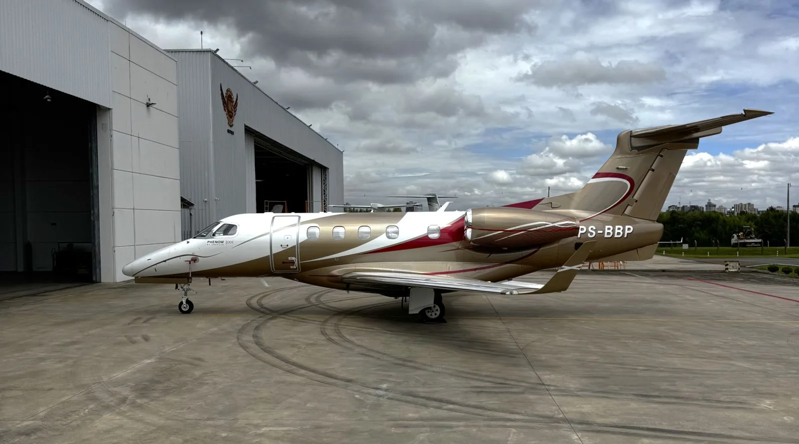 A white, gold, and red Embraer Phenom 300E business jet (PS-BBP) is parked on an airport apron next to a hangar under a cloudy sky.