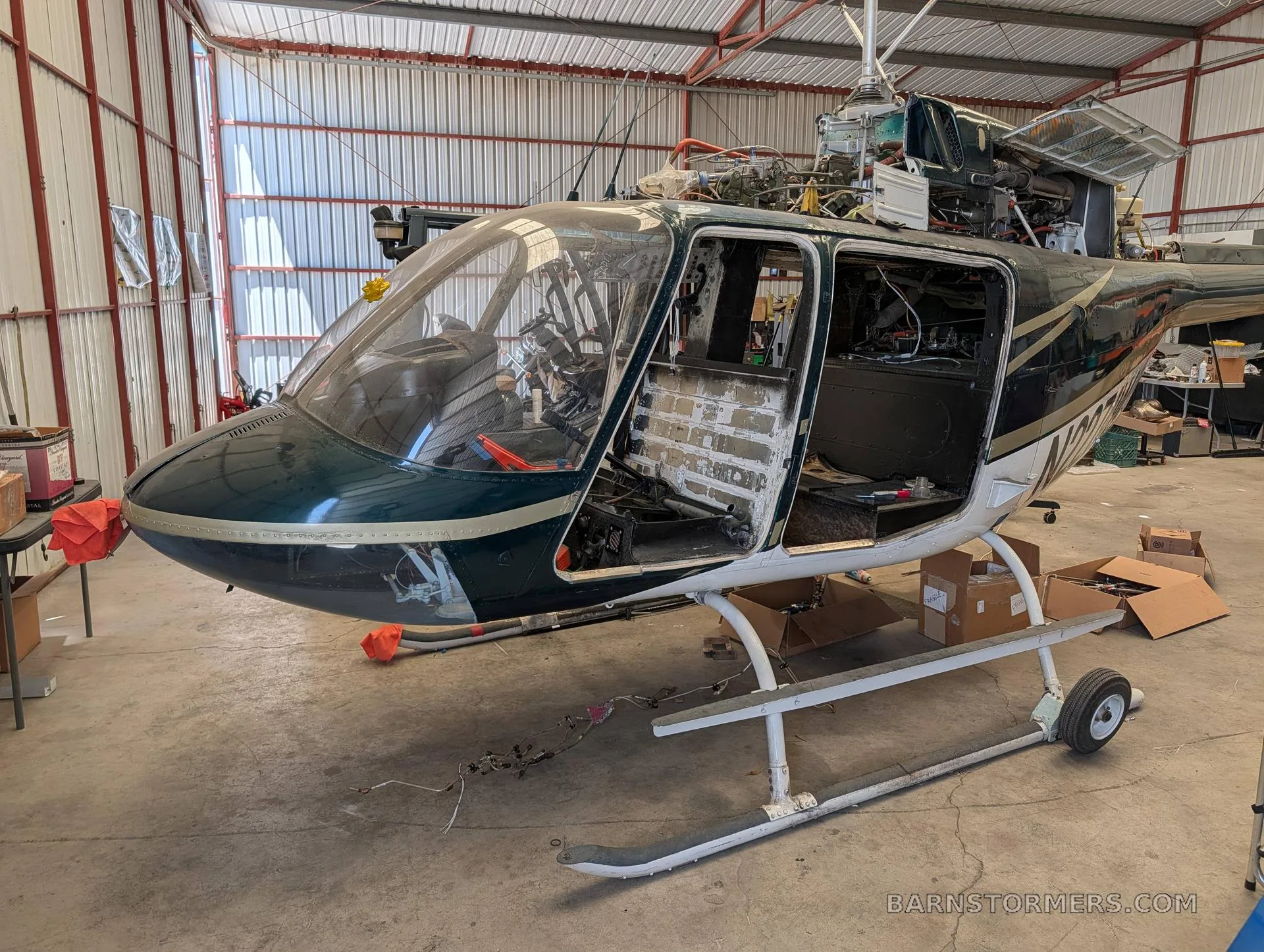 A dark green and white Bell 206 JetRanger helicopter, partially disassembled with its doors open, sits in a hangar.